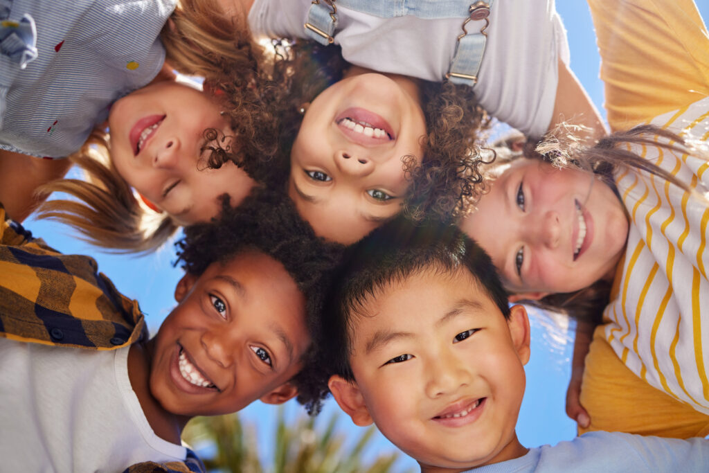 Group of smiling children in a huddle – A group of diverse children looking up and smiling while huddling together, enjoying each other's company under a bright sky.