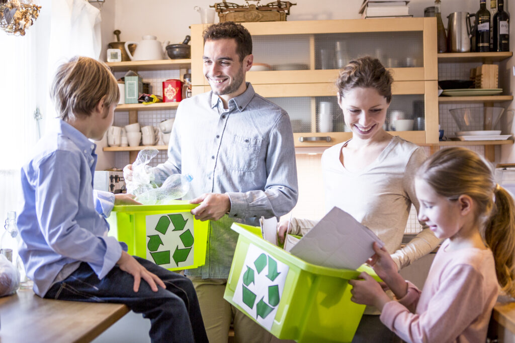 Family teaching children how to recycle at home, sorting plastic and paper into green recycling bins in the kitchen.