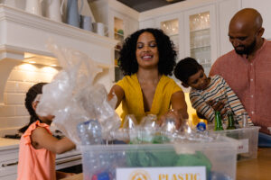 A biracial family sorting plastic and glass bottles for recycling at home. The mother smiles as she handles plastic, while the father assists their young son with glass bottles, teaching sustainable habits.