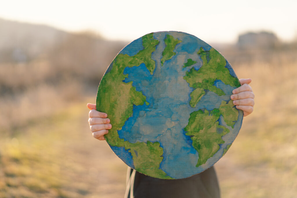 Child holding a globe: A young child holding up a hand-painted, round model of Earth against a blurred outdoor background, symbolizing care for the planet.