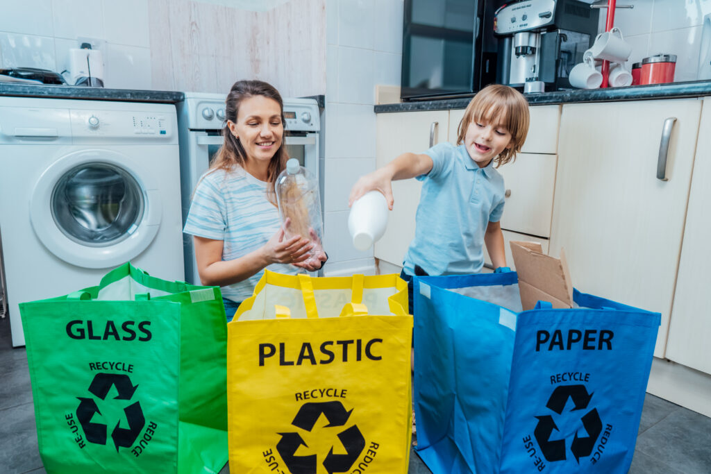 Mother teaching her child to recycle plastic bottles, sorting items into labeled bags for glass, plastic, and paper.