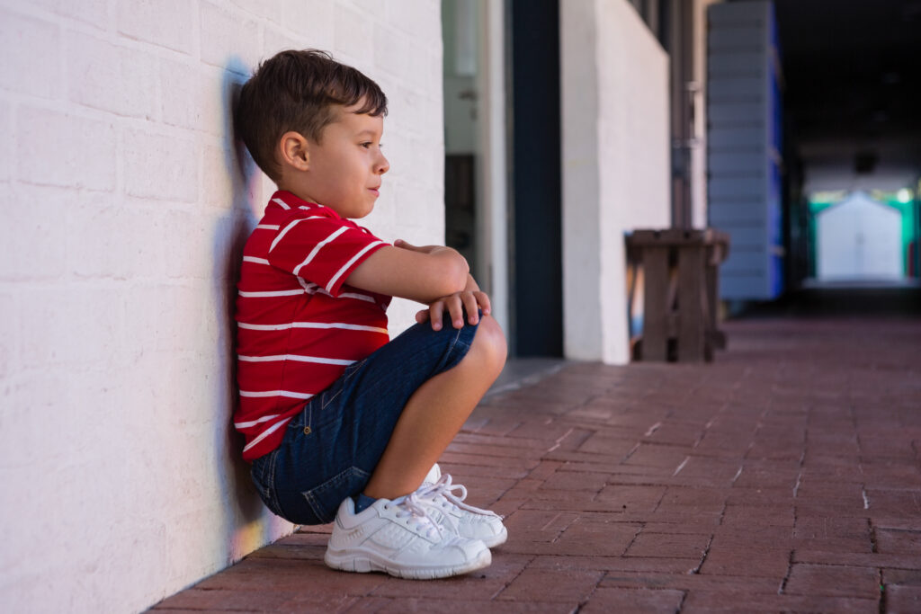 A young boy in a red striped shirt and blue shorts sitting against a white brick wall, looking thoughtful and isolated.