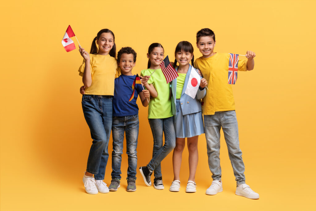 Diverse children holding flags of different countries – A group of children from different ethnic backgrounds holding flags from various nations, standing in front of a vibrant yellow background.