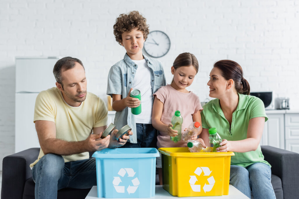 Family sorting recyclable materials into bins, teaching children about recycling cans and plastic bottles.