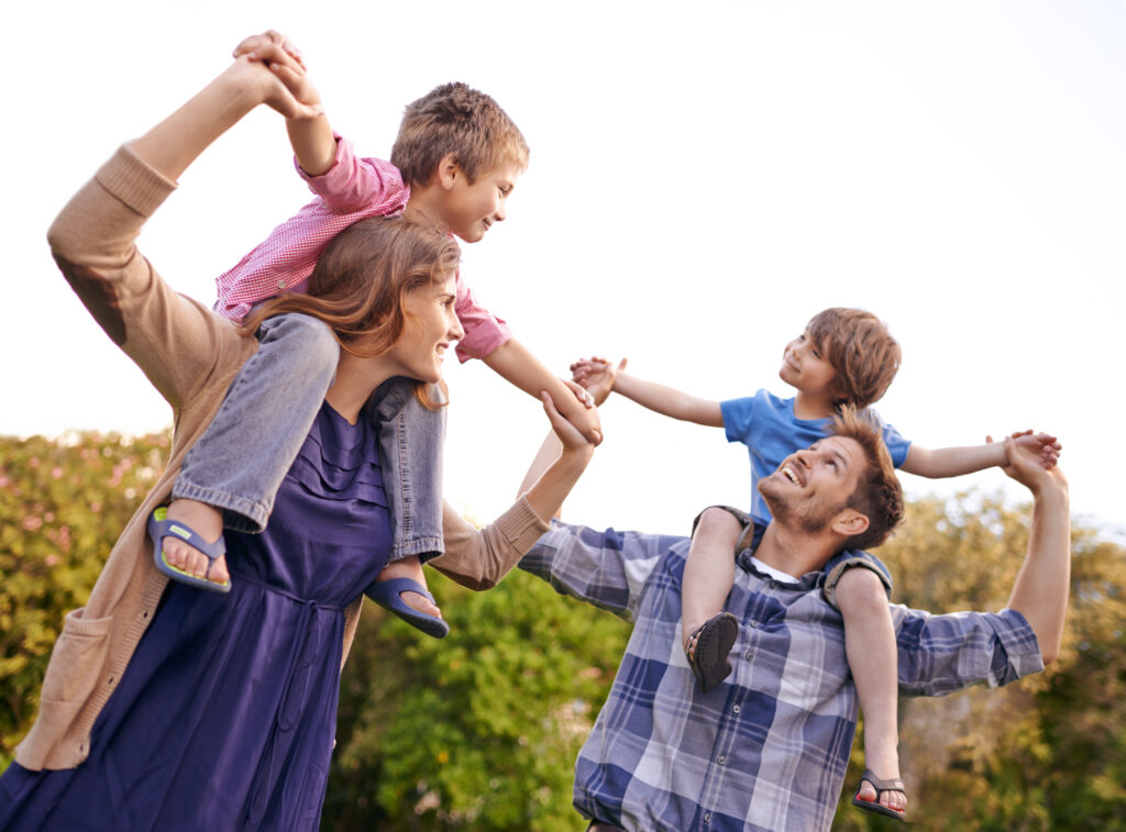 Family enjoying a playful day together, with parents carrying their children on their shoulders outdoors.