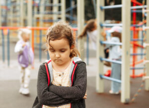 A young girl feeling socially isolated, standing alone at a playground while other children play, highlighting the theme of childhood isolation.